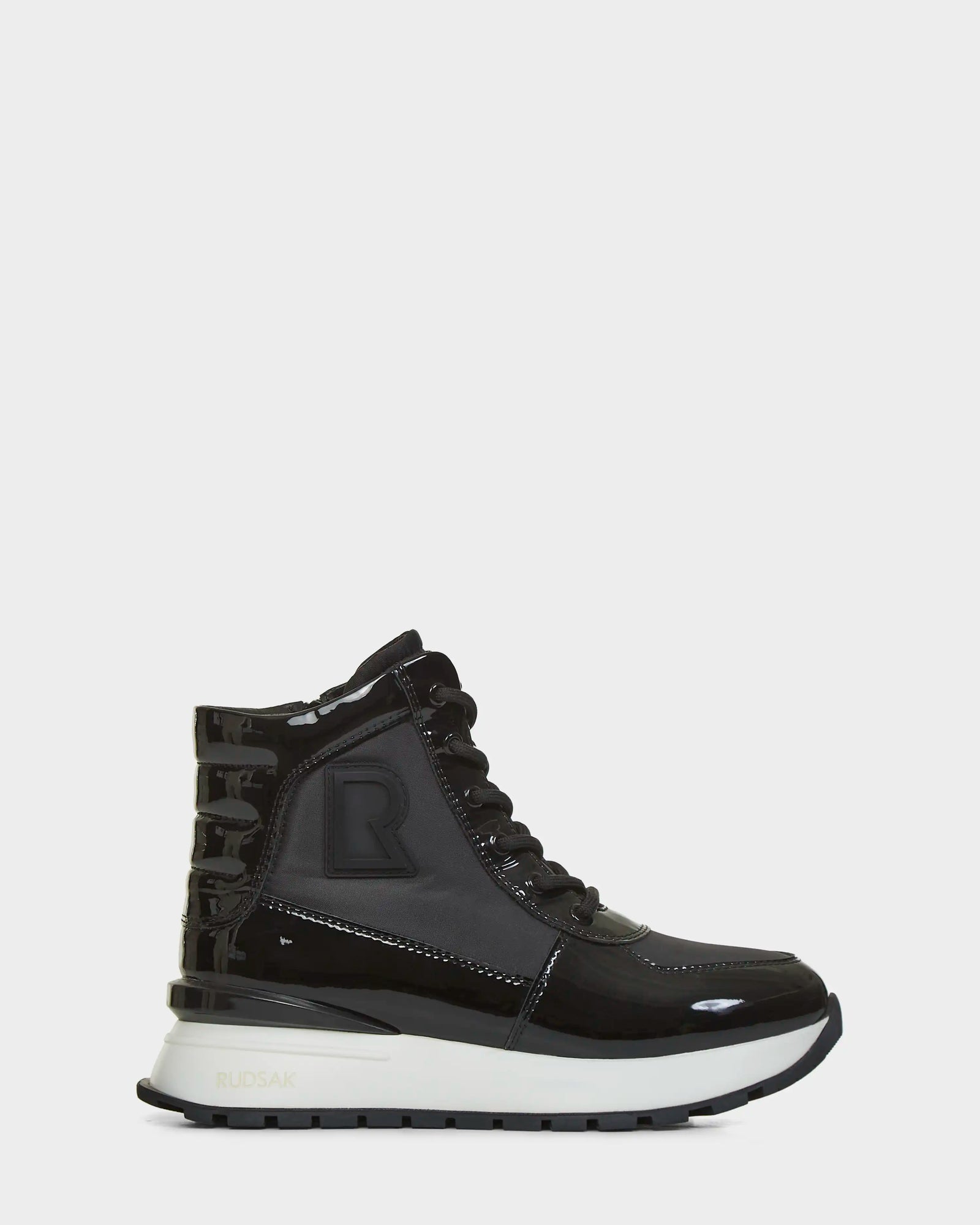 The CHELSA - L223505 Black by Rudsak is a chic black high-top sneaker featuring shiny patent panels, black laces, a thick white and black sole, and a bold raised R logo—offering women comfort and sporty style.