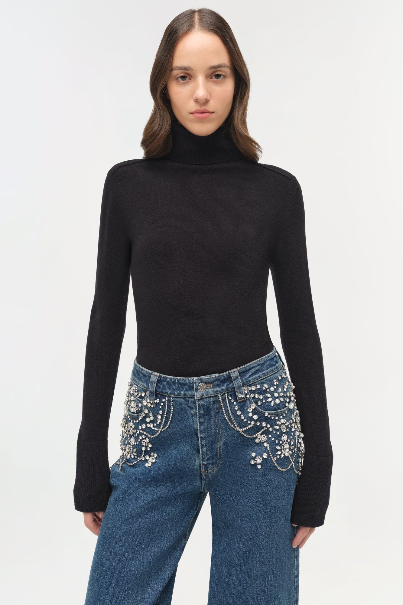 Blue denim crop jeans with rhinestone embellishments on the pockets and waistband, worn with a black turtleneck.