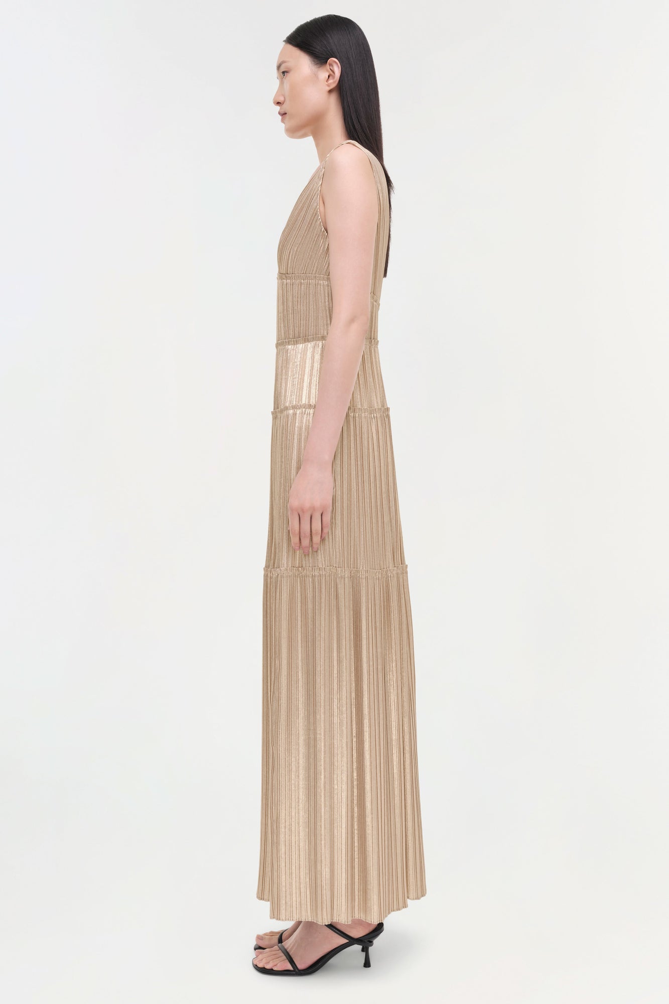 Beige pleated sleeveless gown with vertical striping and tiered panels, shown on model from side profile.