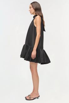 Black sleeveless mini dress with bow neckline and tiered ruffle hem, shown on model in side profile.