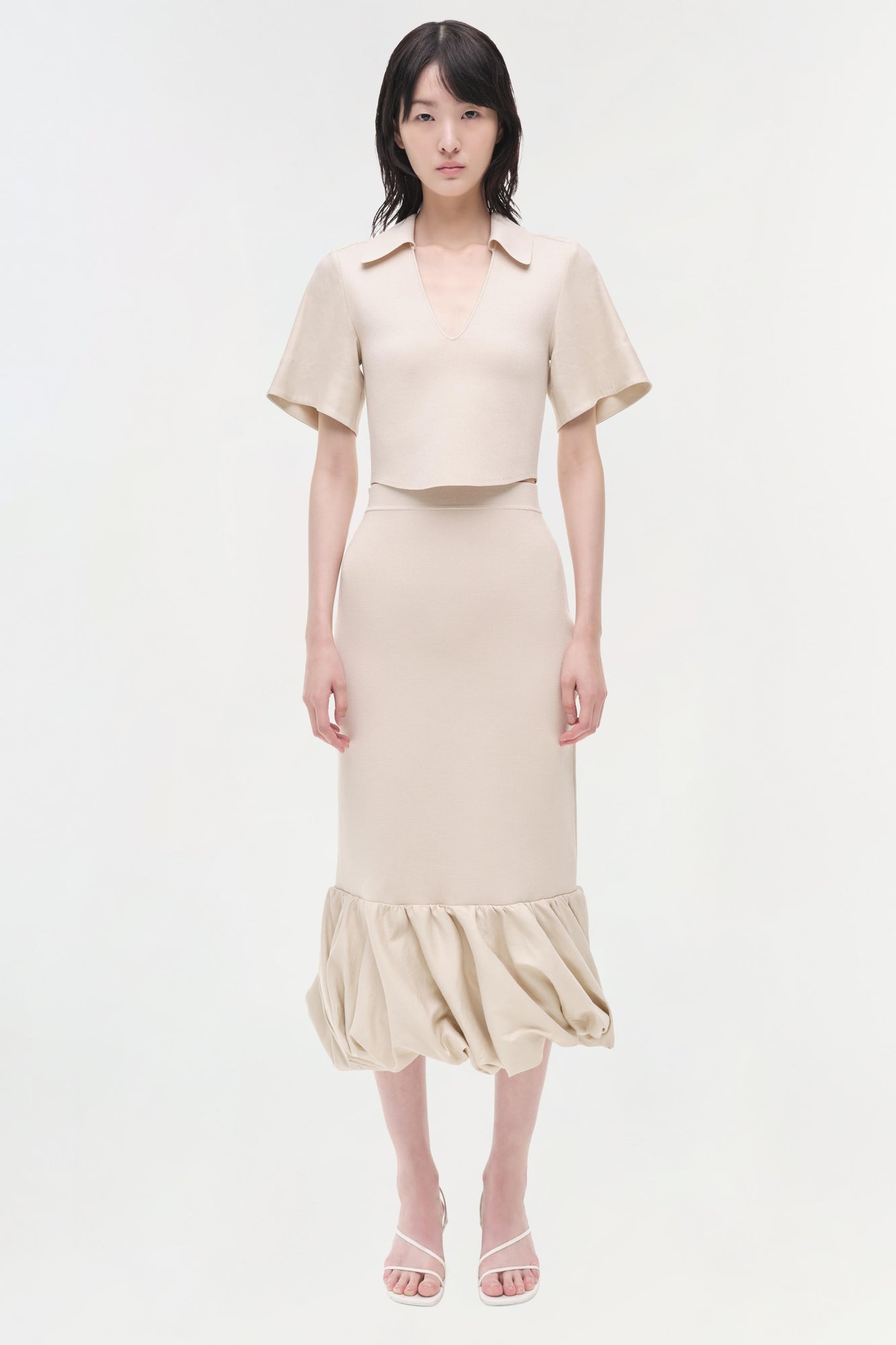 Beige linen knit midi skirt with gathered hem, worn with matching short-sleeve crop top and pink sandals.