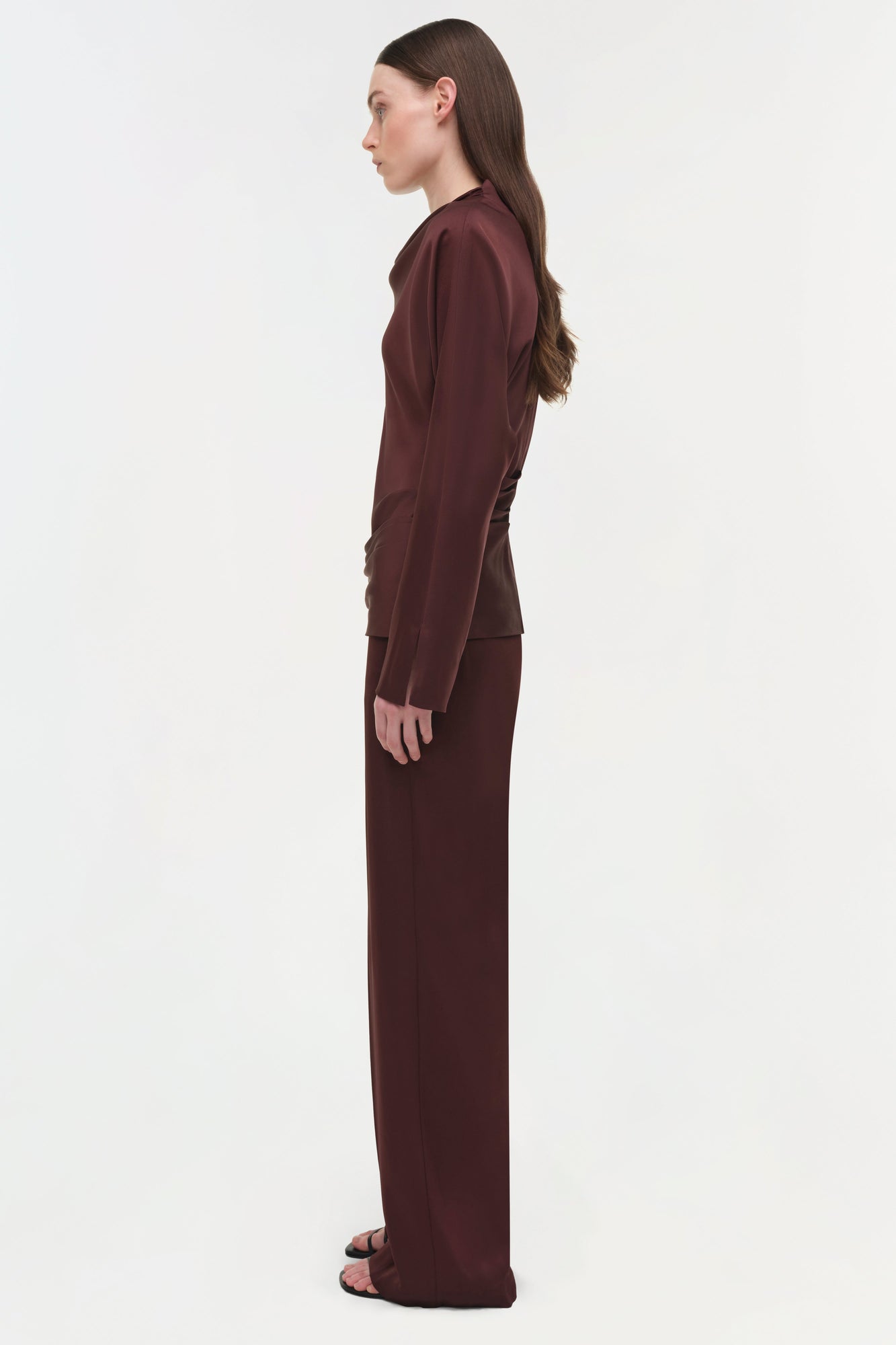 Burgundy satin long-sleeve top with draped fabric detail, shown on model in side profile.