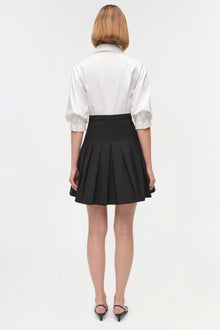 Woman wearing white collared shirt with half-length sleeves and black pleated mini skirt, viewed from back.