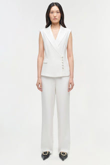Woman wearing white sleeveless blazer vest and straight leg trousers with button details.