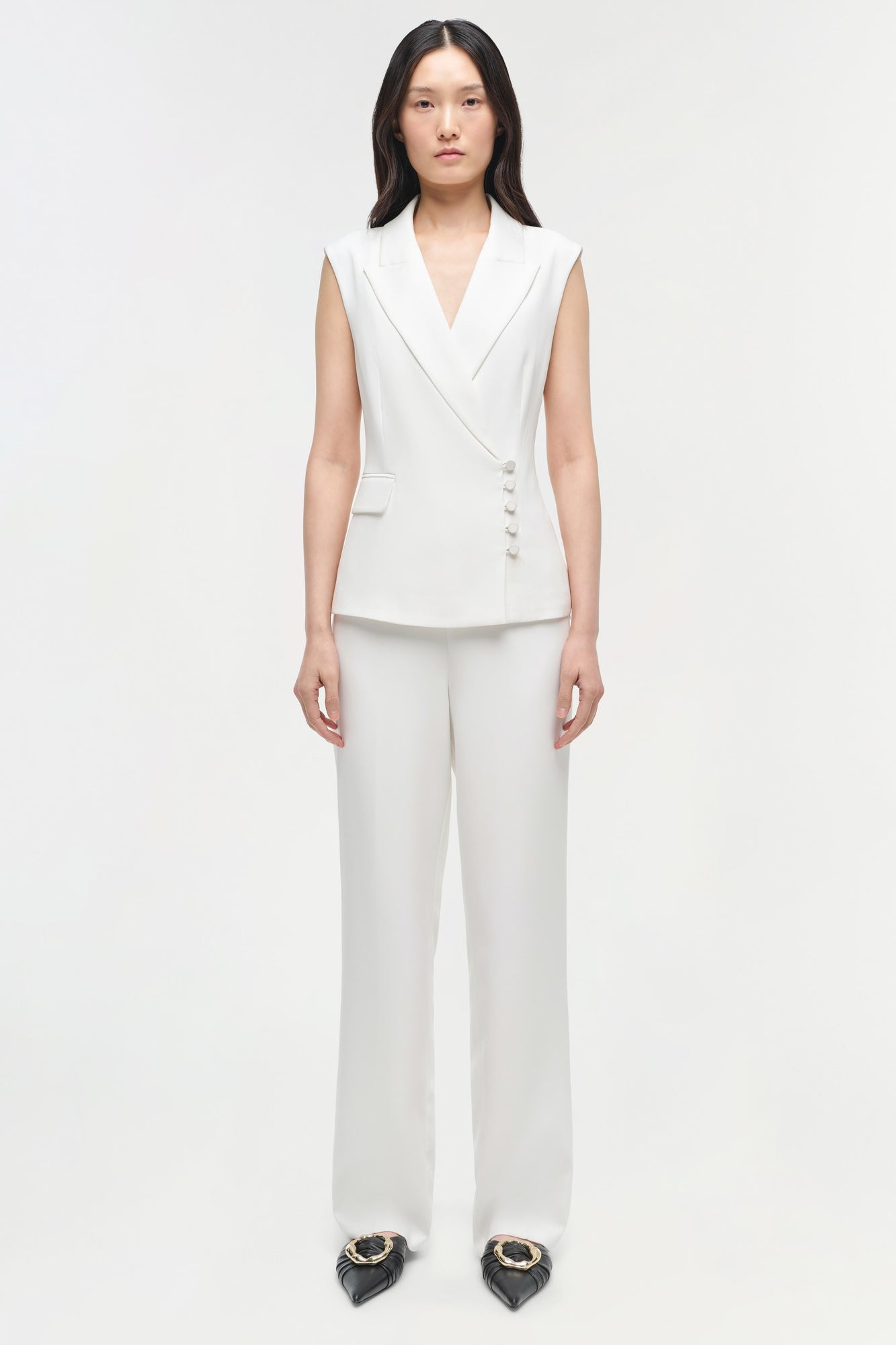 Woman wearing white sleeveless blazer vest and straight leg trousers with button details.