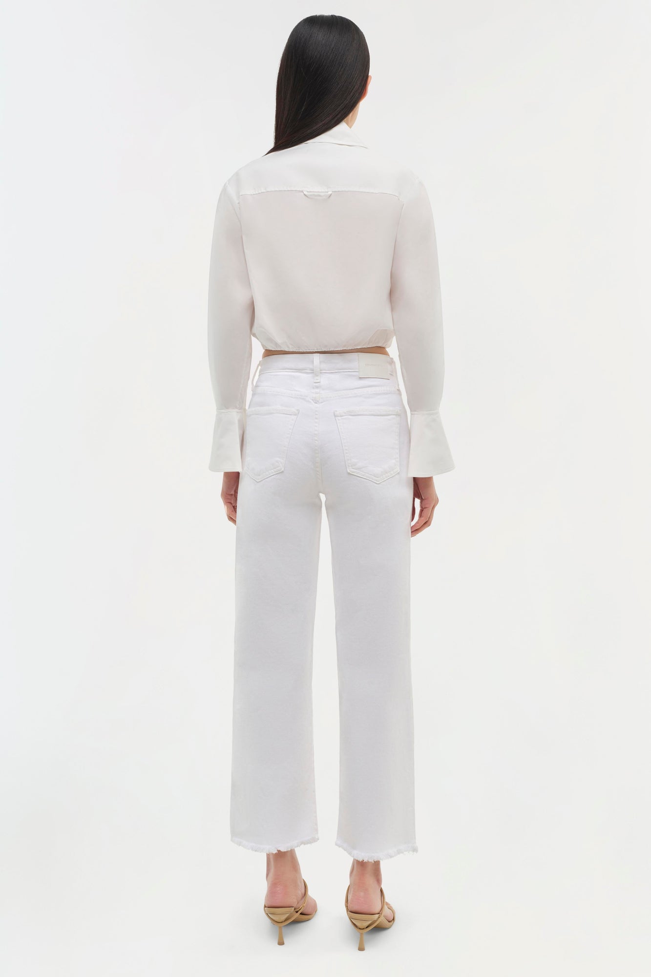 Woman wearing cream cropped long-sleeve top with flared cuffs and white cropped jeans, shown from back.