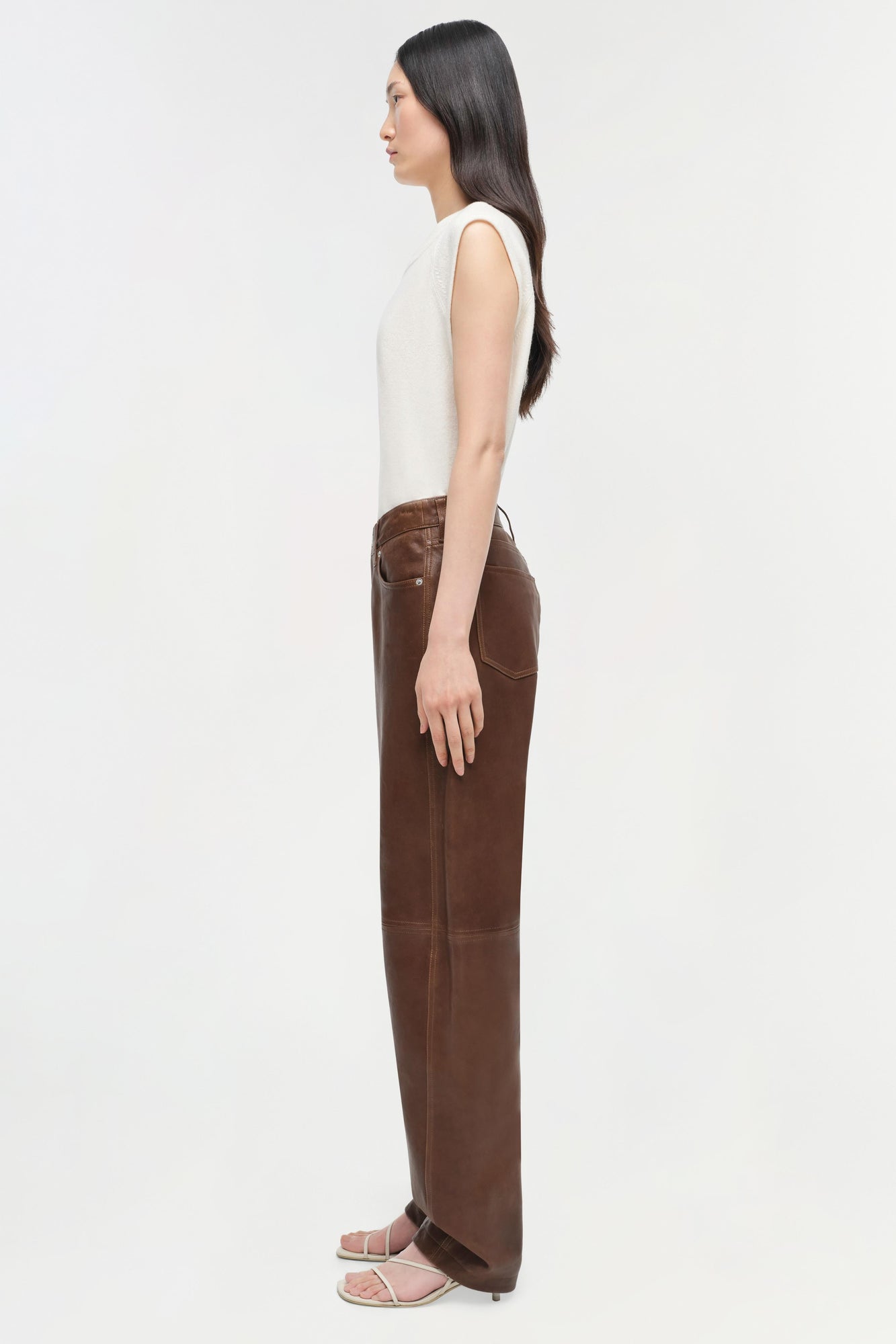 Woman wearing cream sleeveless cashmere vest over brown trousers, shown in side profile.