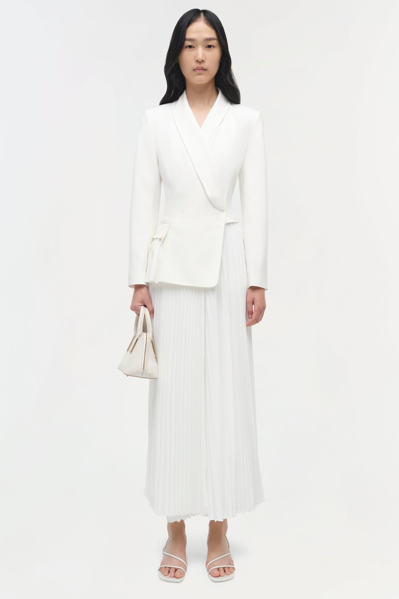 Woman wearing cream pleated blazer midi dress with long sleeves, paired with white sandals and matching handbag.
