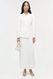 Woman wearing a cream-colored blazer with wrap front paired with a pleated midi skirt and white sandals.
