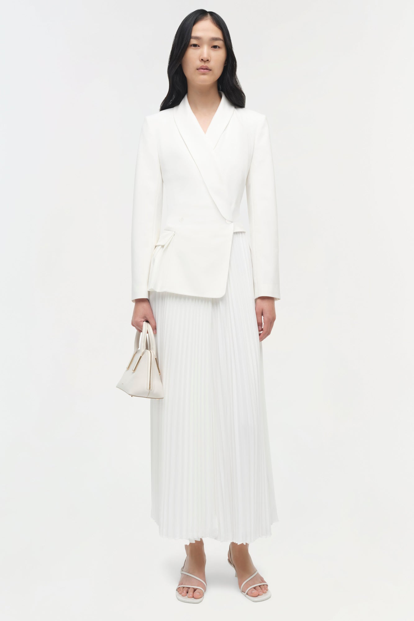 Woman wearing a cream-colored blazer with wrap front paired with a pleated midi skirt and white sandals.