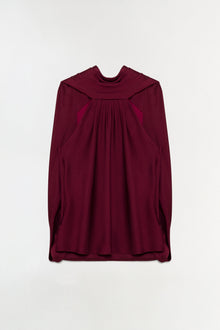 Deep burgundy satin cape top with draped front panel and long sleeves on neutral background.