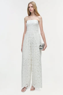 Woman wearing white lace strapless jumpsuit with wide legs and metallic clutch.