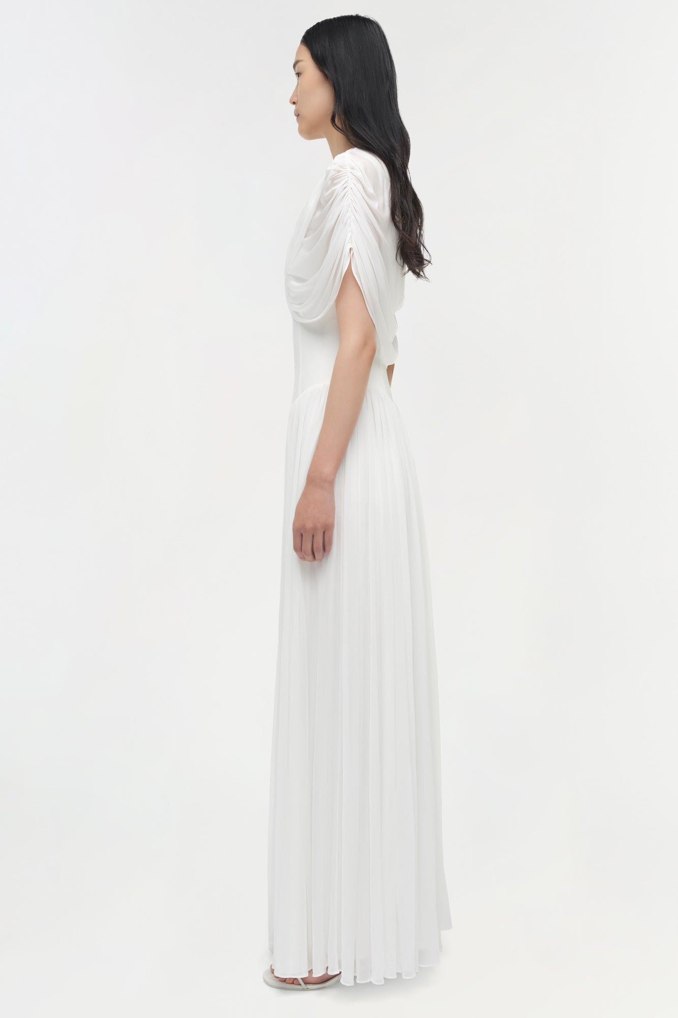 White draped jersey cape maxi dress with pleated skirt and side cutout detail, shown on model in profile.