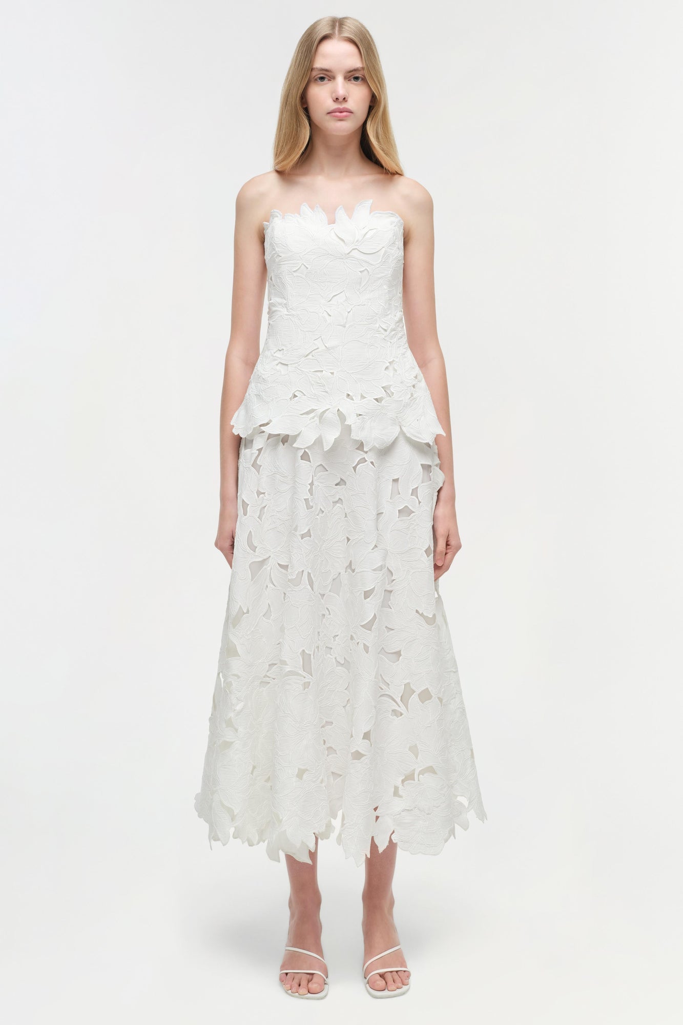 White strapless midi dress with leaf-shaped embroidered cutout details throughout, worn with white sandals.
