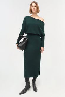 Woman wearing dark green off-shoulder midi dress with long sleeves and a fitted waist, paired with black boots and a black ba