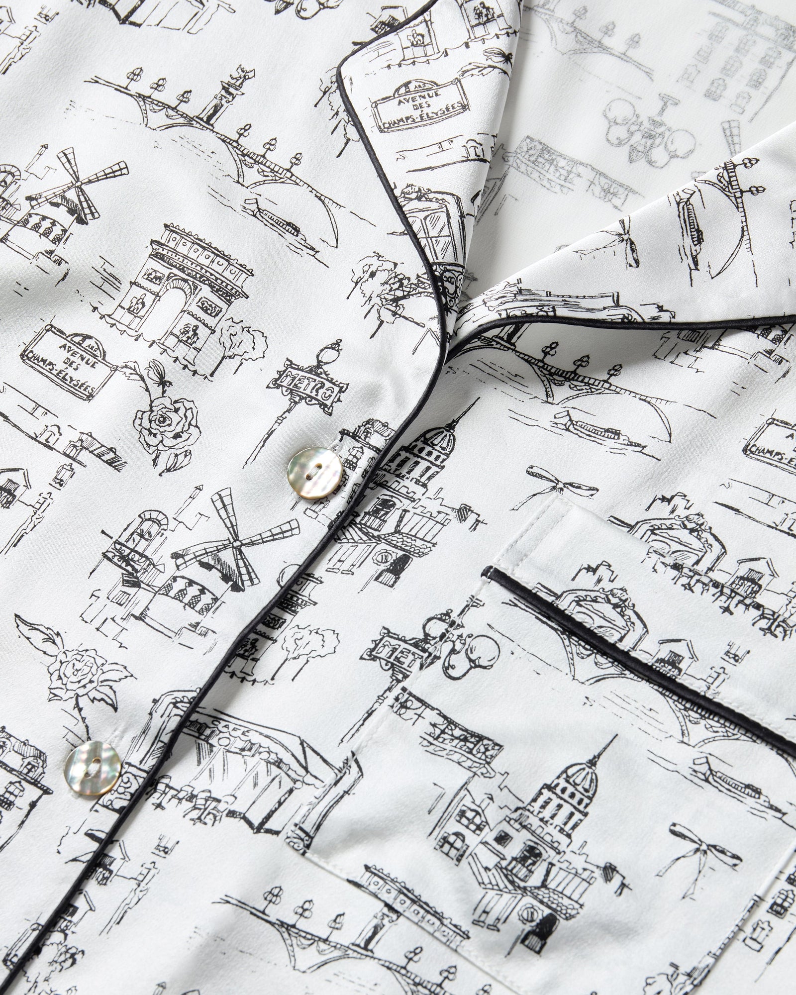 Women's Silk Pajama Set in Parisian Toile: white button-up with black piping, illustrated with buildings and trees. Includes long-sleeve top with chest pocket and matching long pants featuring an elastic waistband.
