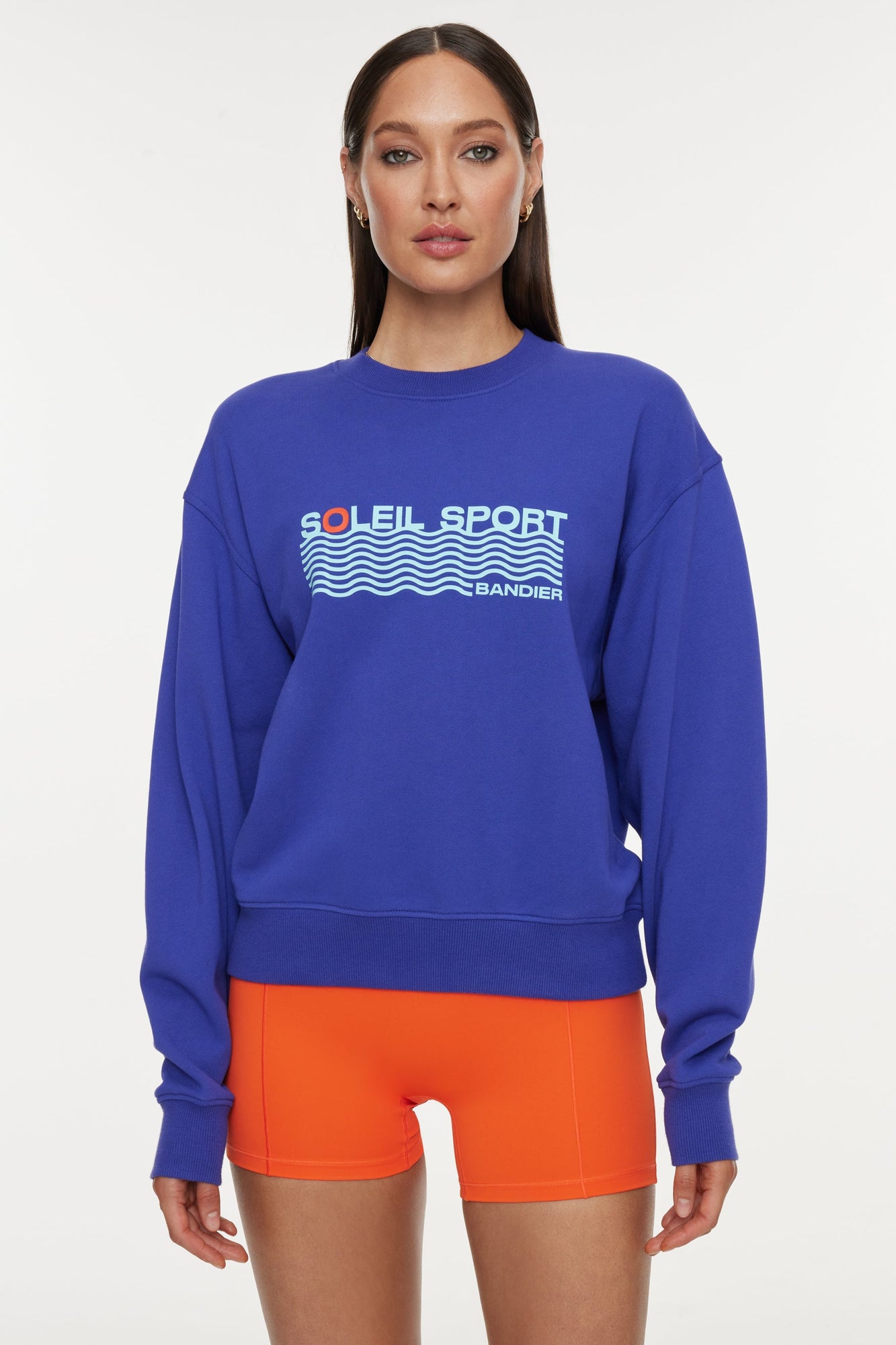 Model wears a royal blue crewneck sweatshirt with the lettering "Soleil Sport" across the chest with wavey lines below and "Bandier" in the bottom right corner. 