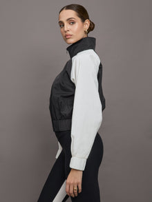 Model wears a cropped windbreaker with a popped collar, black bodice and white long sleeves with a cinched wristline.