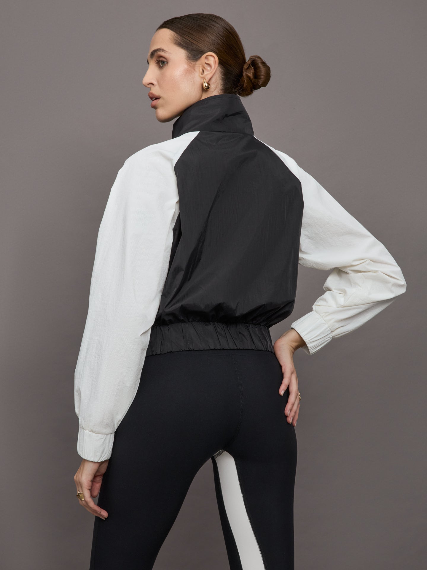 Model wears a cropped windbreaker with a popped collar, black bodice and white long sleeves with a cinched wristline.