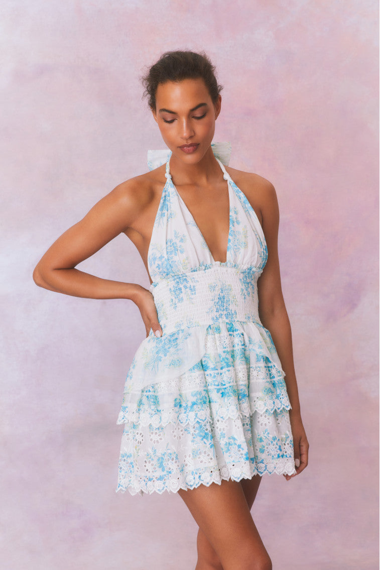 White halter dress with a blue floral pattern, halter ties in bow at neck and below, is a wide smocked waistband and the skirt has two ruffled tiers.