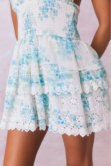 White halter dress with a blue floral pattern, halter ties in bow at neck and below, is a wide smocked waistband and the skirt has two ruffled tiers.