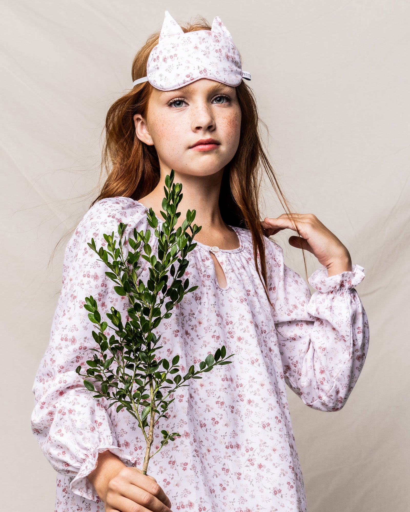A young red-haired girl wears Petite Plumes Kids Kitty Sleep Mask in Dorset Floral. Holding green leaves against a plain backdrop, she embodies the ideal gift for fans of whimsical bedtime accessories.