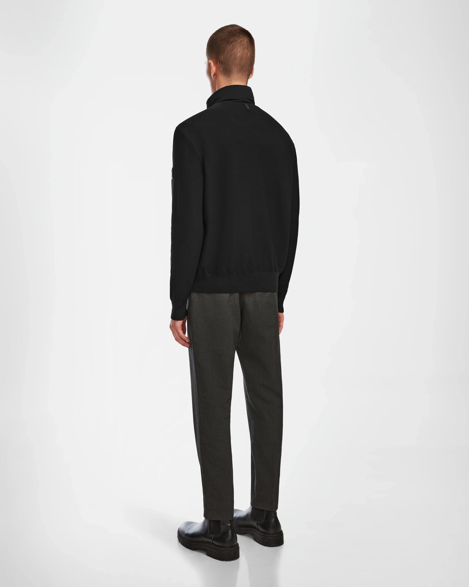 A man stands facing away from the camera, wearing the Rudsak EUGENE - 6124016 Black hybrid jacket, dark gray trousers, and black shoes against a plain white background.