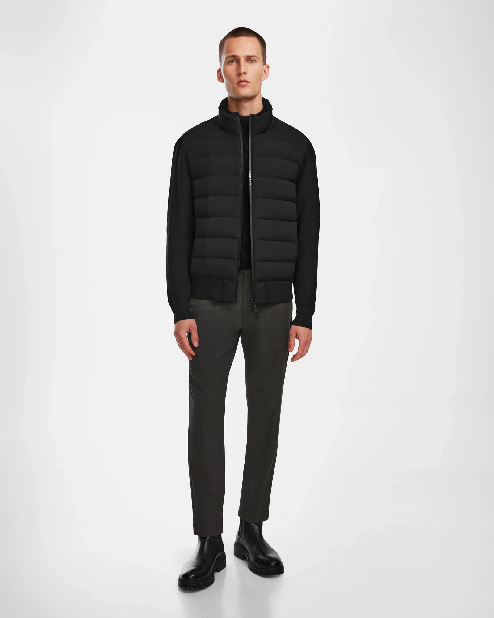 A man stands against a white background, wearing the Rudsak EUGENE - 6124016 Black jacket over a black shirt, paired with dark brown pants and black boots. He faces forward with his arms relaxed at his sides.