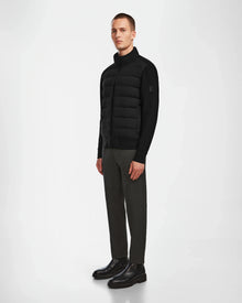 A man stands against a plain white background wearing the Rudsak EUGENE - 6124016 Black mens hybrid jacket, dark gray pants, and black boots. He faces slightly left with his arms relaxed by his sides.