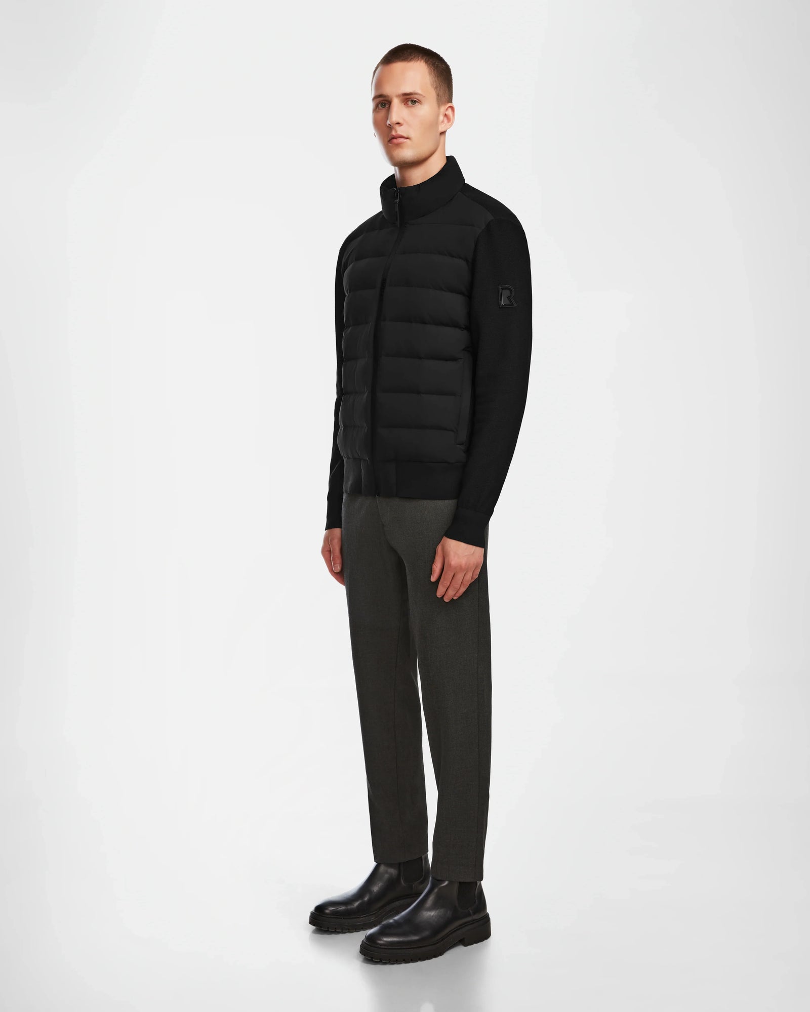 A man stands against a plain white background wearing the Rudsak EUGENE - 6124016 Black mens hybrid jacket, dark gray pants, and black boots. He faces slightly left with his arms relaxed by his sides.