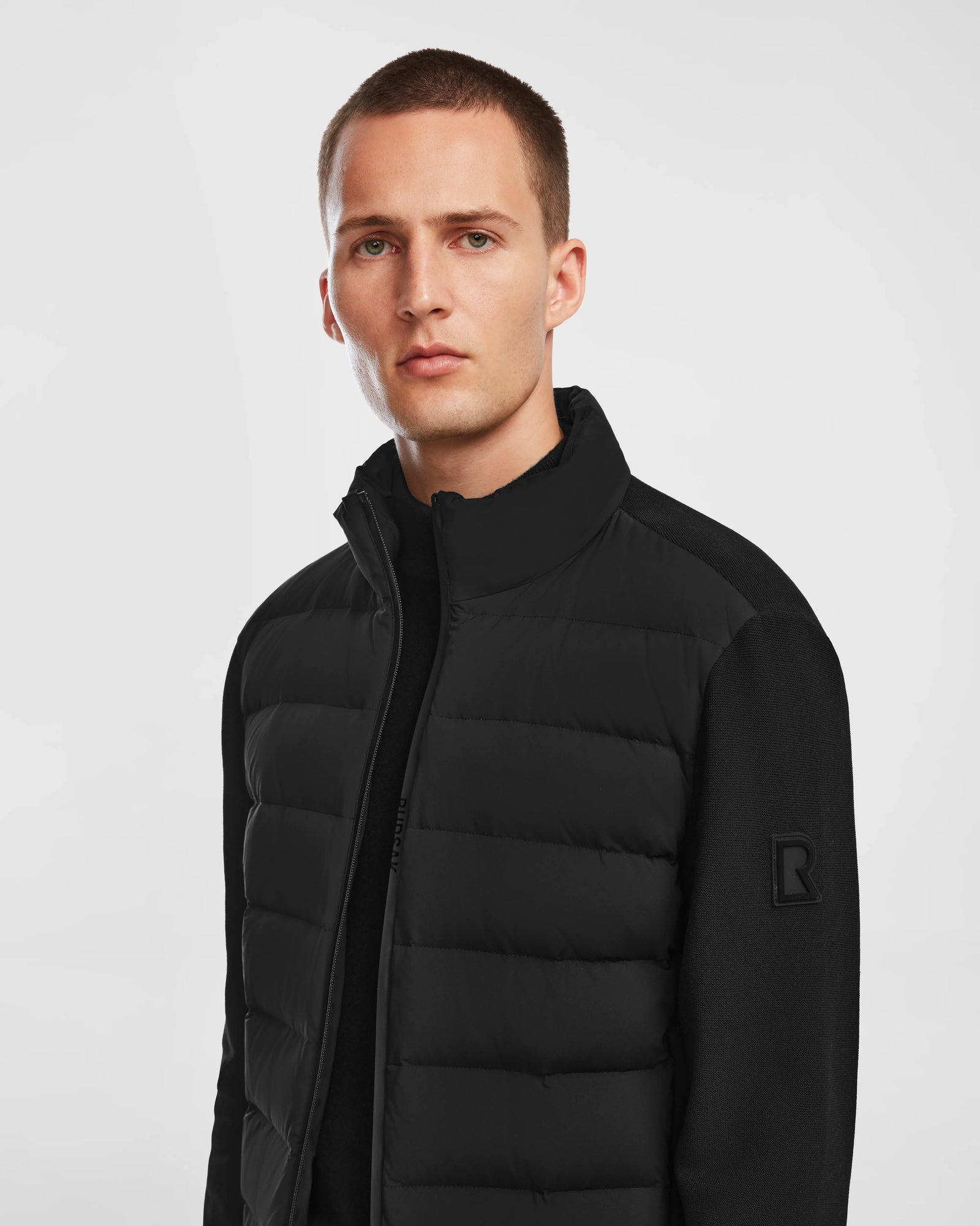 A man with short hair wears the Rudsak EUGENE - 6124016 Black jacket, featuring a high collar and subtle R logo on the sleeve, standing against a plain light background.