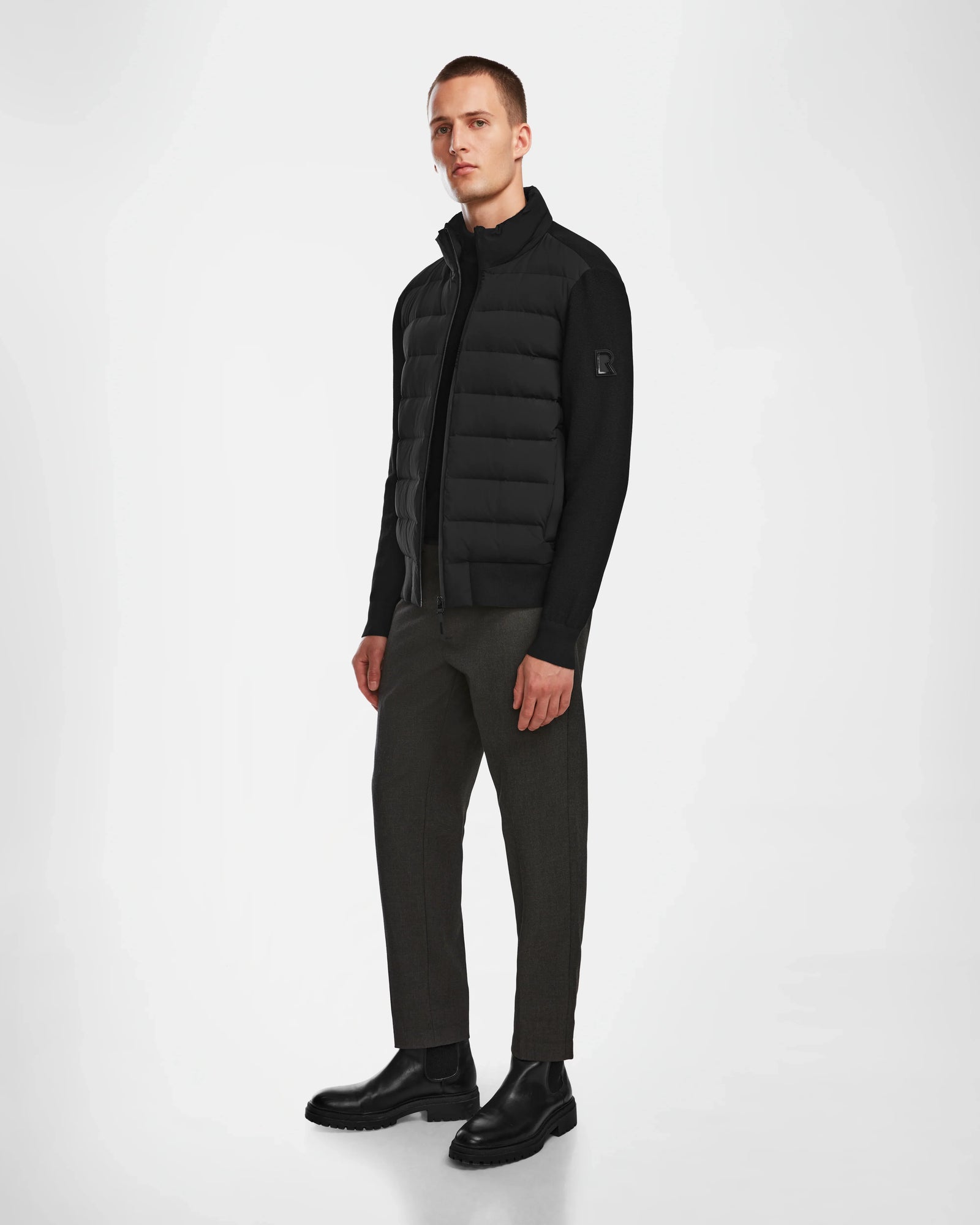 A man stands against a plain white background wearing the Rudsak EUGENE - 6124016 Black mens hybrid jacket, dark pants, and black boots. He faces slightly to the side with a neutral expression and his arms relaxed by his sides.