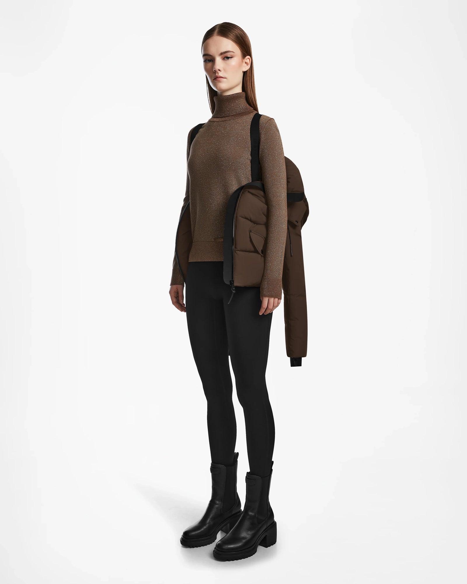 A young woman with straight hair stands against a white background, wearing black leggings and boots, carrying the Rudsak ADELINE - 8124544 Mocha womens down puffer over one shoulder.