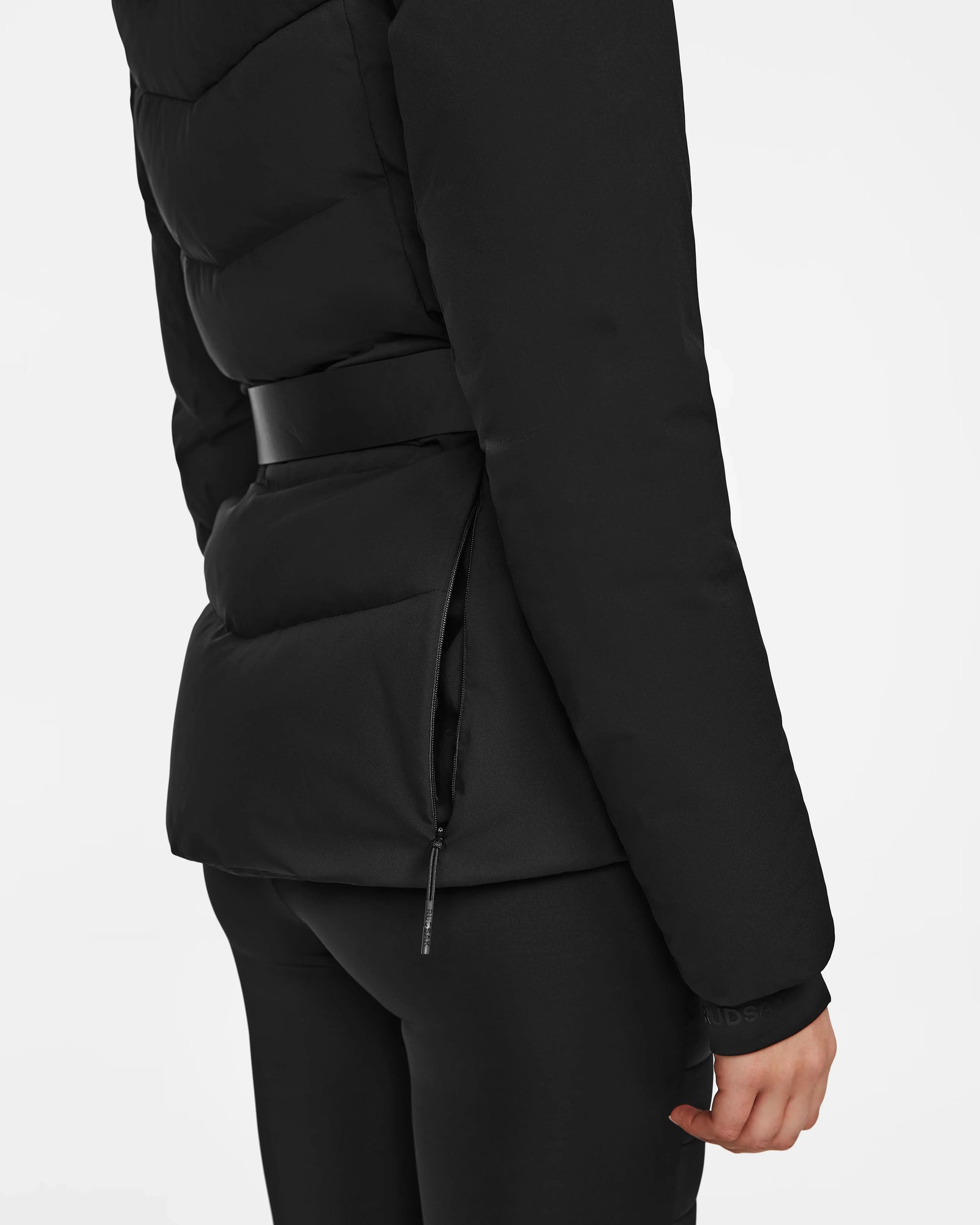 A woman is shown from the back and side wearing the Rudsak ALAIA - 8124816 Black fitted quilted ski jacket with a belted waist, visible zipper pocket, and black pants; only her torso and arm are visible.
