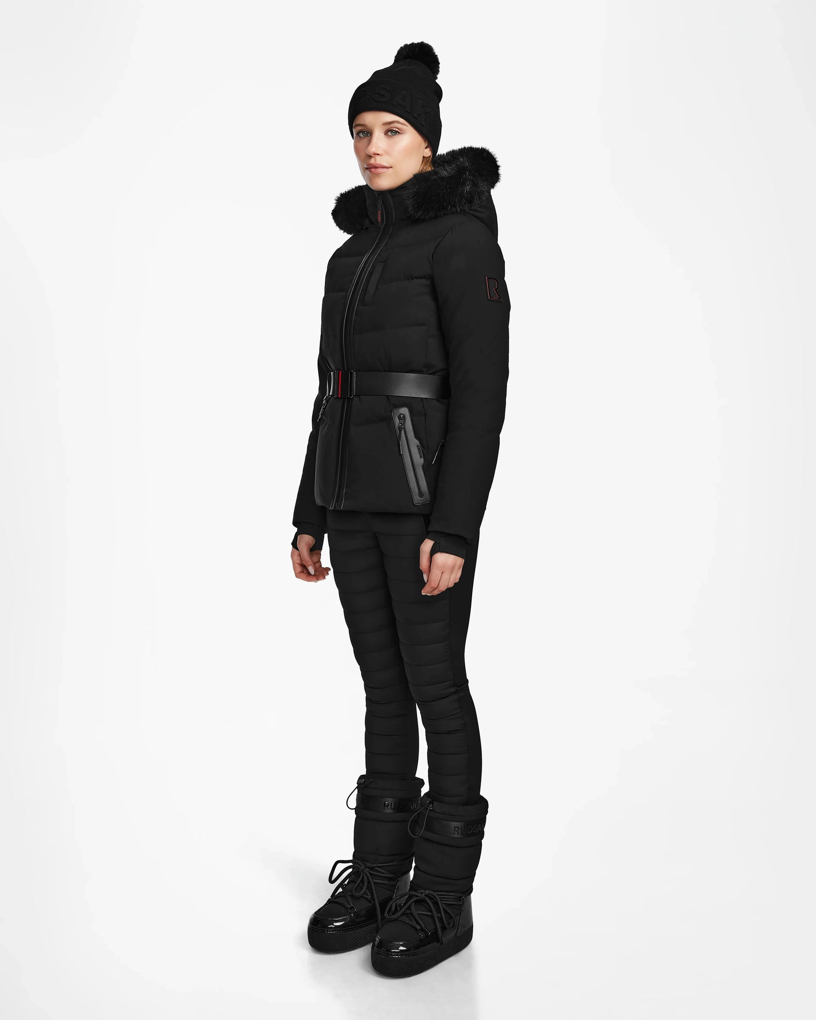 A woman wears the Rudsak ALAIA - 8124816 Black womens quilted ski jacket with a fur-trimmed hood, belted puffer design, matching pants, black beanie, and winter boots—ideal for the slopes—against a white background.