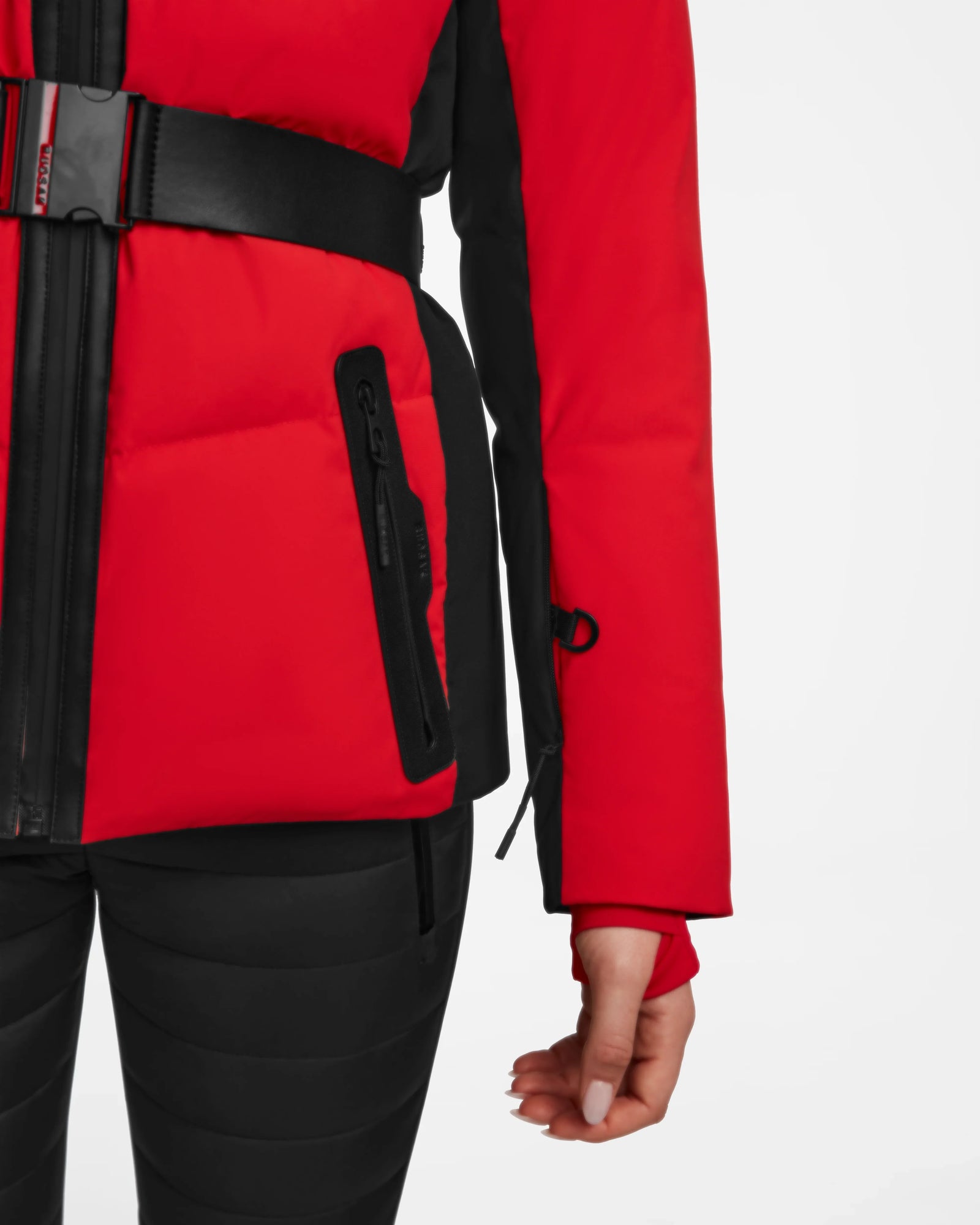 A person wears the Rudsak ALAIA - 8124816 Flame Red ski jacket, featuring red and black colors, a black belt, and zippered pocket, paired with black pants. Only the lower torso and right arm appear against a white background.