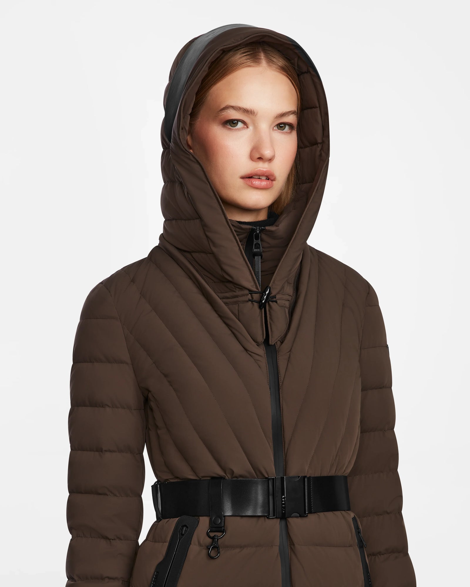 A woman wears the Rudsak ALISA - L124506 Espresso quilted down puffer winter coat with a black vegan leather belt and zipper, standing against a plain, light background.
