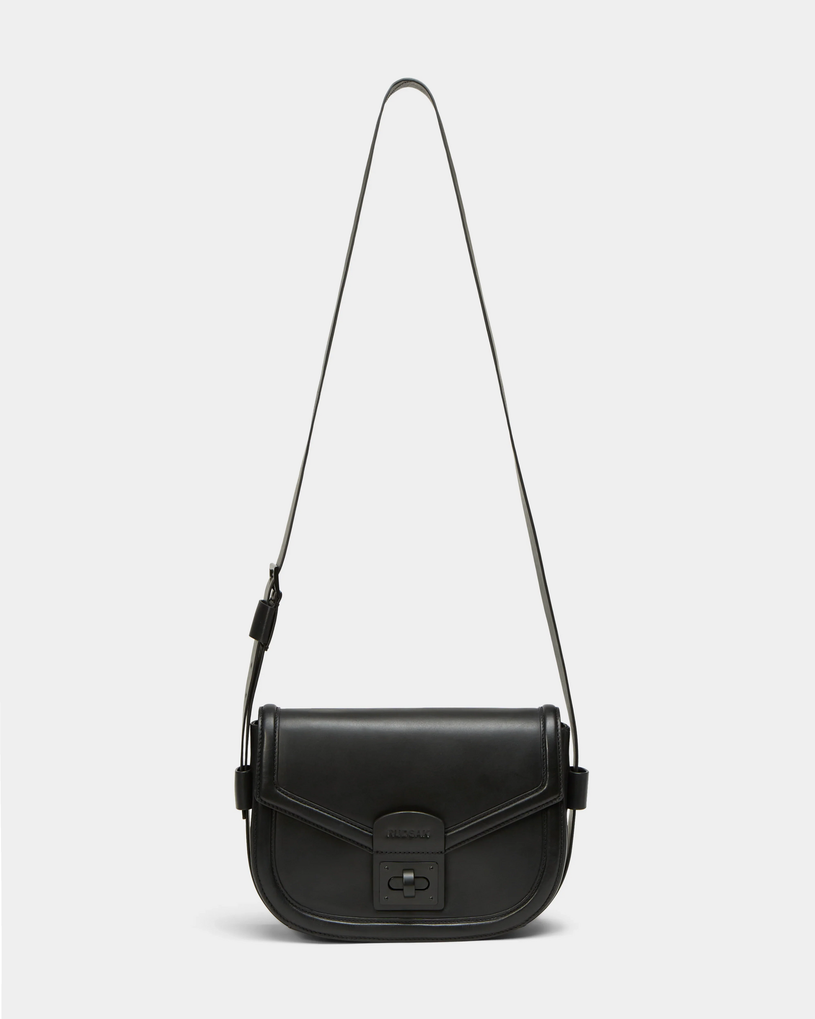 The Rudsak ALORA - 8324506 is a minimalist black crossbody bag crafted from premium leather, with a long adjustable strap, flap closure, and front clasp. Shown here against a plain white background.