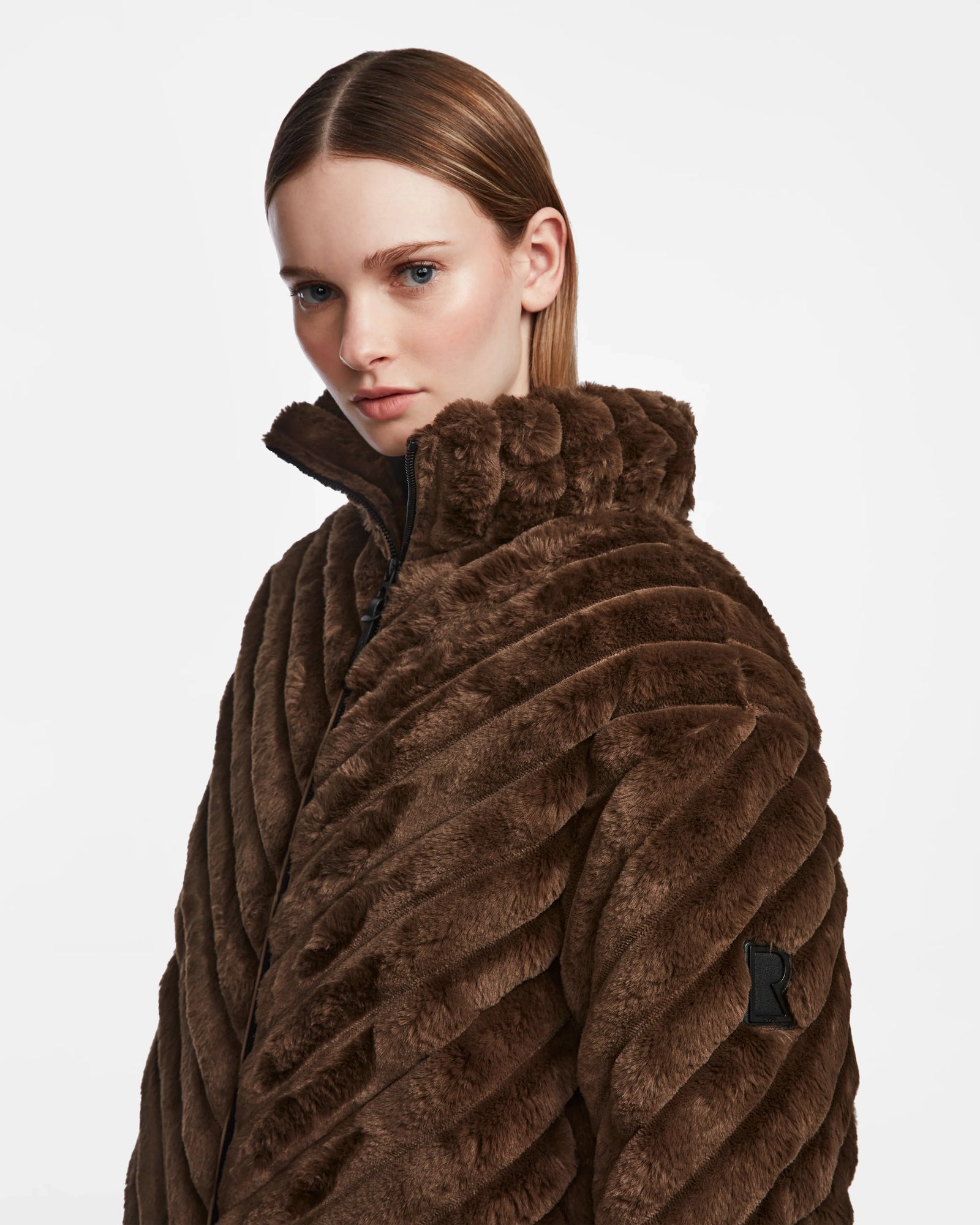 A woman with straight, light brown hair models the Rudsak ANASTASIA - 8124804 Espresso textured dark brown winter jacket with a high collar, standing against a plain white background.