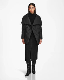 A woman faces forward against a white background, wearing the Rudsak ANNE MARIE - 8124713 Black mid-length jacket with an oversized collar, paired with a black turtleneck, long front-slit skirt, and knee-high boots.