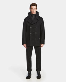 A man faces forward wearing the Rudsak ARTHUR - 6124521 Black premium wool blend double-breasted peacoat with a hood, paired with black pants and boots, against a plain white background.