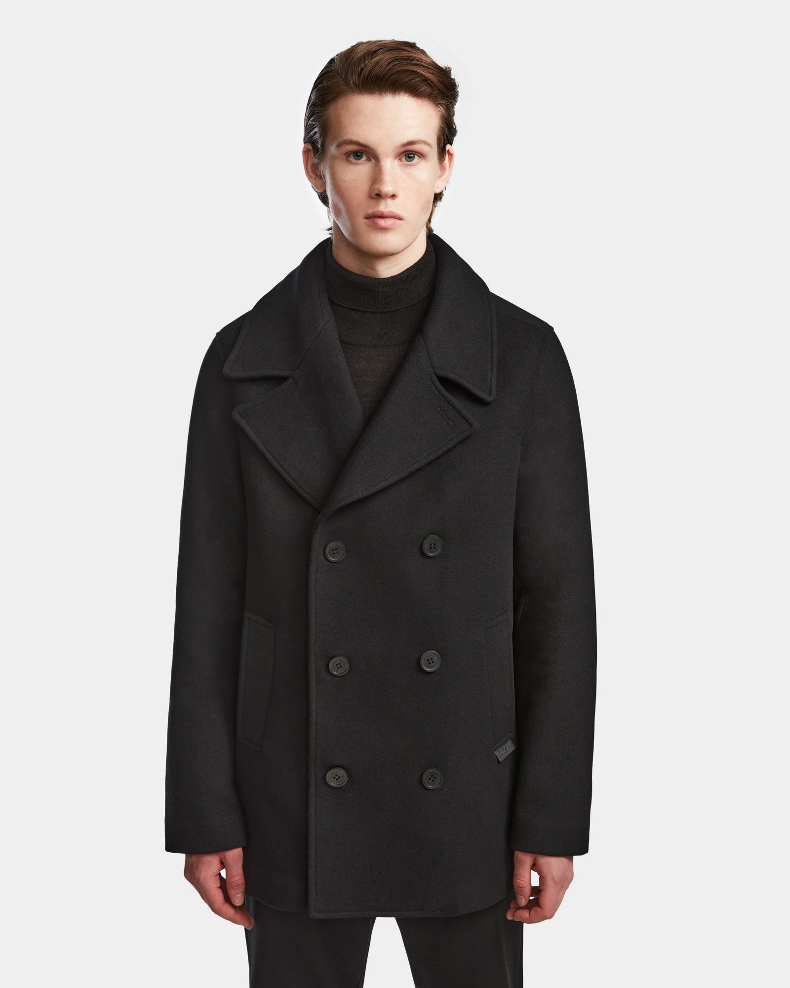 A person with short brown hair wears the Rudsak ARTHUR - 6124521 Black mens wool peacoat, featuring a black double-breasted design over a black turtleneck, standing against a plain white background and facing forward neutrally.