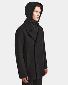A man models the Rudsak ARTHUR - 6124521 Black double-breasted peacoat with large lapels and a hood, made from premium wool blend, standing against a plain white background and gazing slightly to the side.