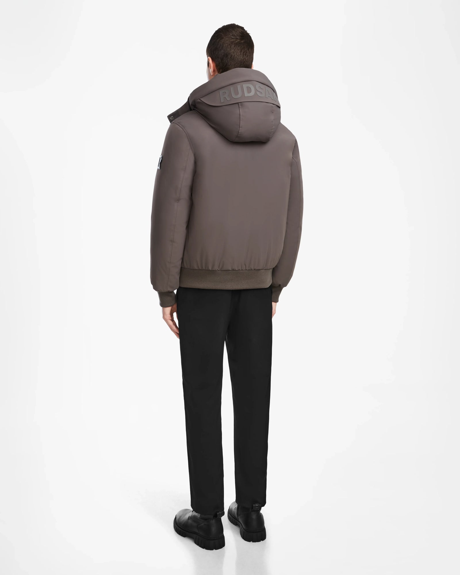A person stands facing away, wearing the Rudsak ASH - 6124599 Grey water-resistant bomber with RUDSAK on the hood, black pants, and black shoes against a plain white background.