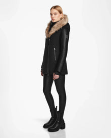 A woman wears the Rudsak AURORE R - L124535 Black down parka with vegan leather sleeves, paired with black leggings and chunky boots, standing against a white background and facing slightly to the side with a neutral expression.