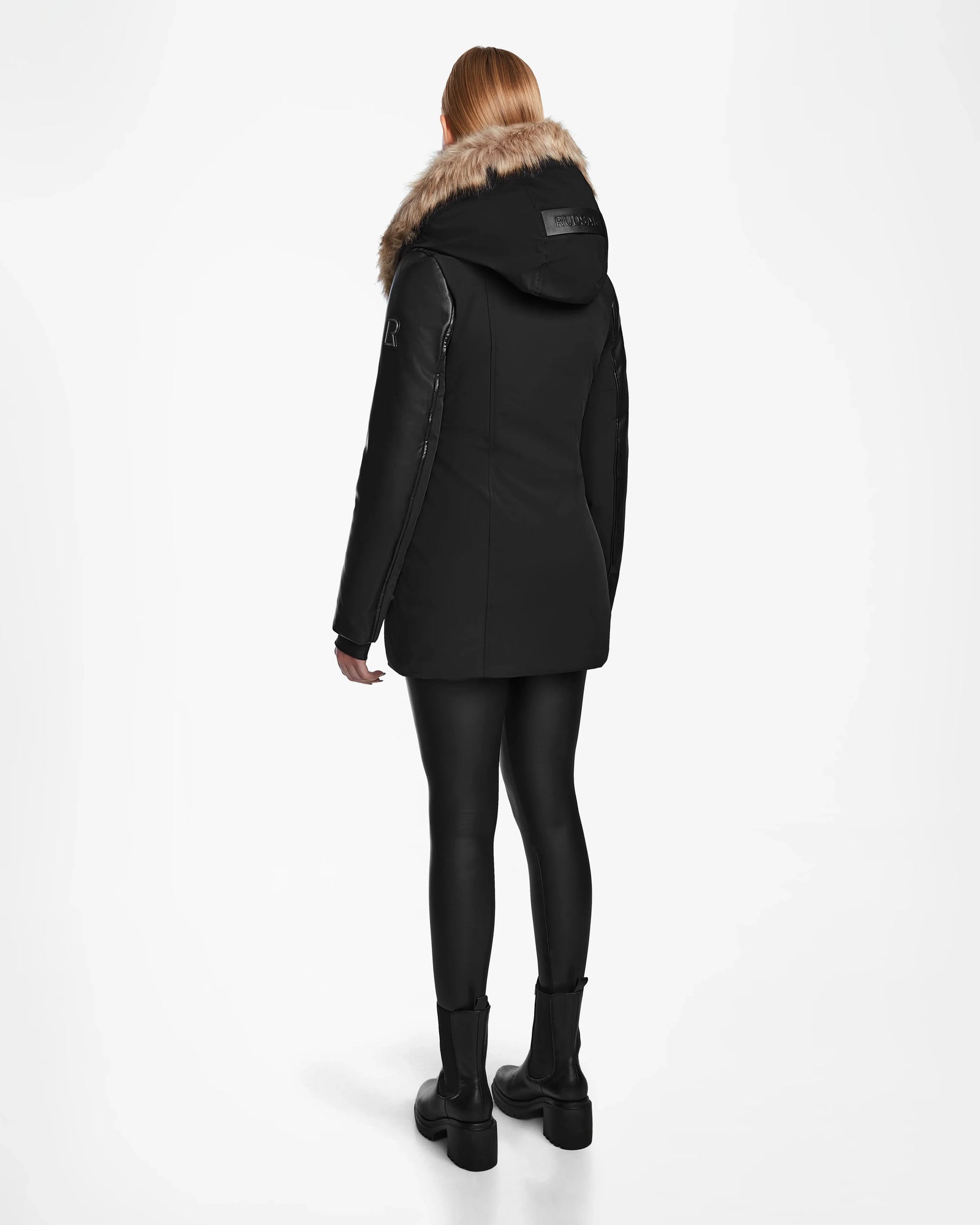 A person with long, light hair, seen from the back, wears Rudsak's AURORE R - L124535 Black weather-resistant jacket with faux fur hood, black leggings, and black boots against a plain white background.