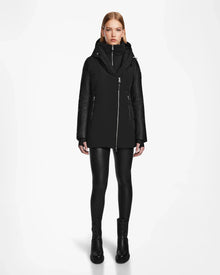 A woman faces forward against a white background, wearing the Rudsak AURORE R - L124535 Black weather-resistant jacket with vegan leather sleeves, paired with black leather-look leggings and black boots.