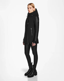 A woman stands against a white background wearing the Rudsak AURORE R - L124535 Black jacket with vegan leather sleeves, paired with black leggings and boots, looking slightly to the side with her arms relaxed.