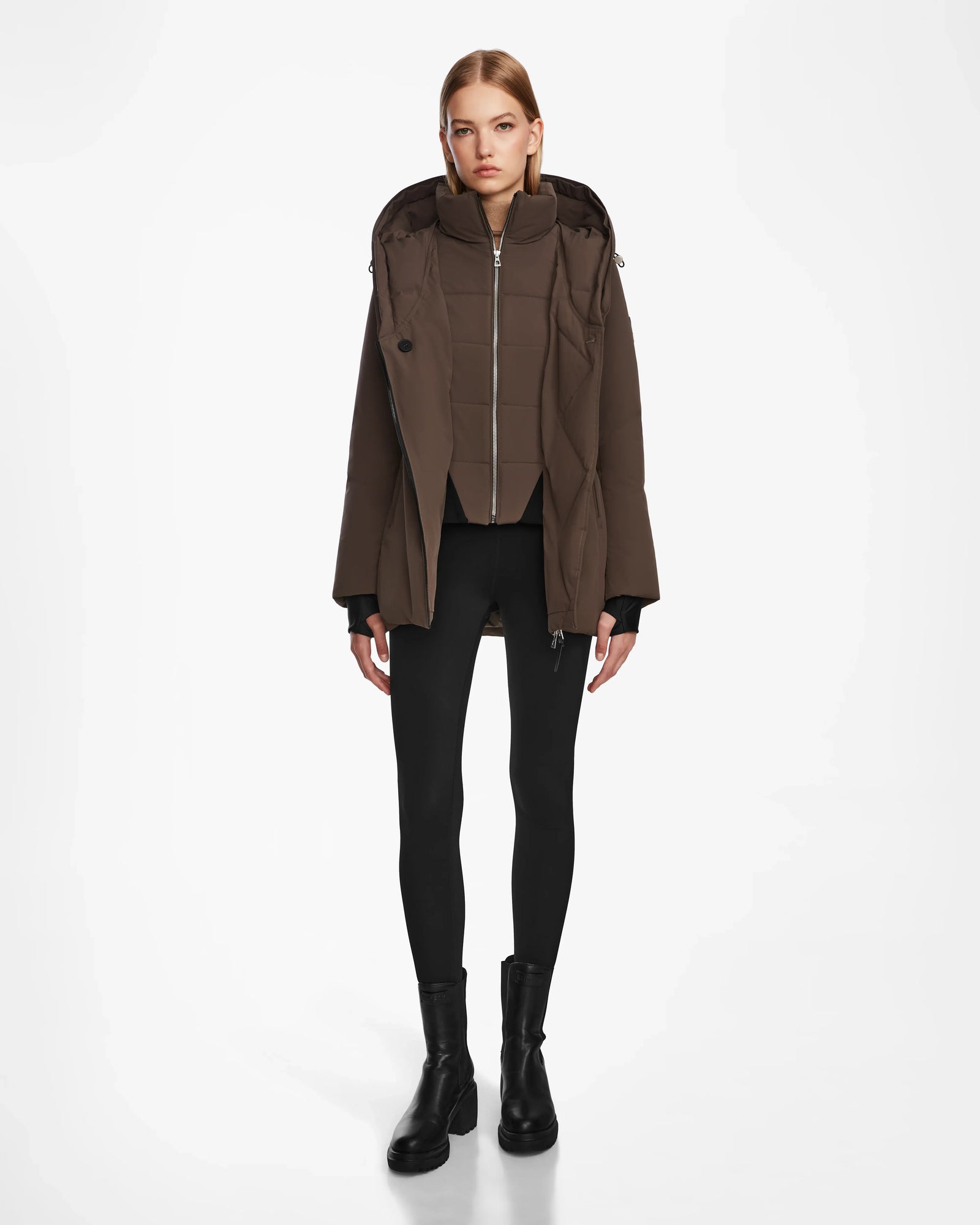 A woman faces forward against a white background, wearing the Rudsak AURORE - L123561 Espresso fitted down parka, paired with black leggings and black ankle boots.