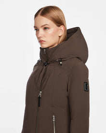 A woman with straight blonde hair wears the Rudsak AURORE - L123561 Espresso, a fitted brown down parka with zippered pockets and a black logo patch on the sleeve, standing against a plain white background.
